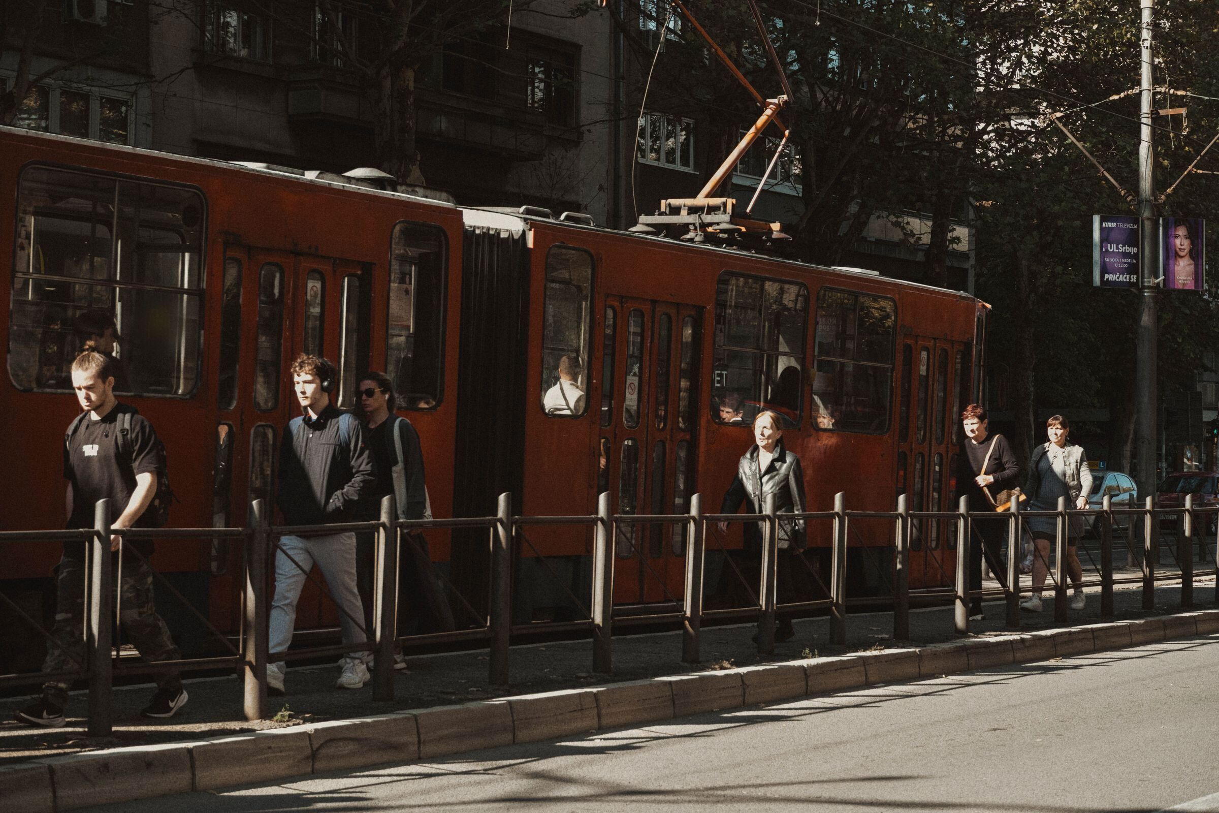 Ljudi sa rancima i torbama idu trotoarom pored crvenog tramvaja, po sunčanom danu.